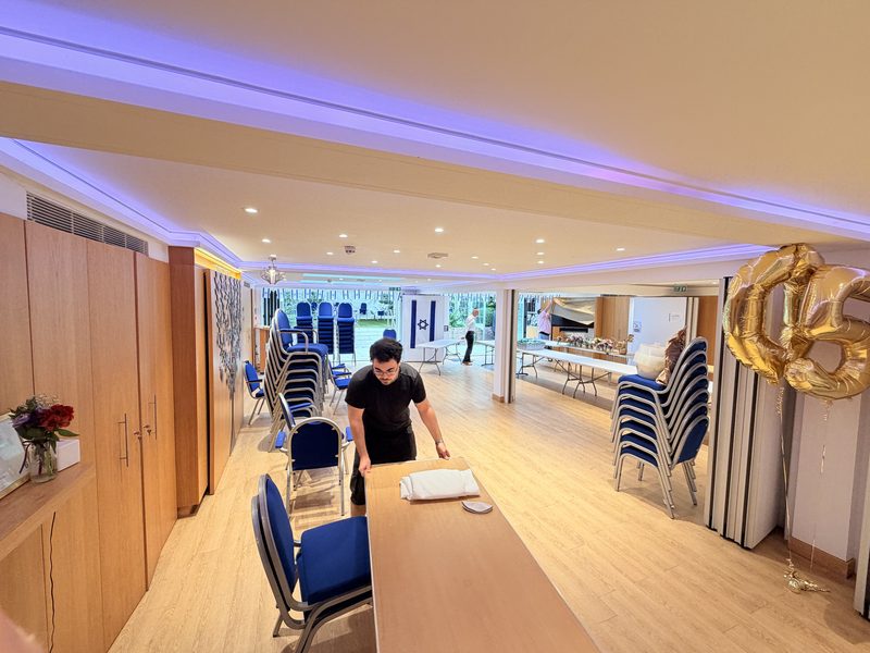 The hall at Hadley Wood Shul being set up for an event, with long tables, stacked blue chairs and the garden marquee beyond