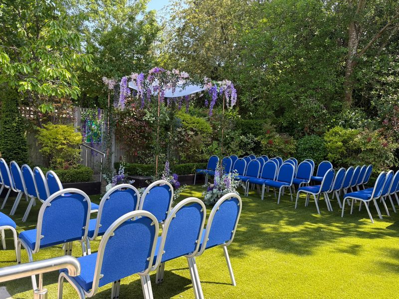 A chuppah decorated with purple flowers set up for a wedding in the garden at Hadley Wood Shul, with rows of blue chairs on the lawn